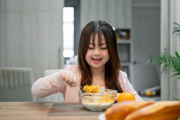 A young adorable and happy Asian girl is enjoying eating a bowl of cereal at a dining table