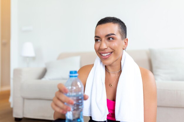 Young adult African American woman drinking water from plastic bottle sitting on fitness mat and resting after training at home