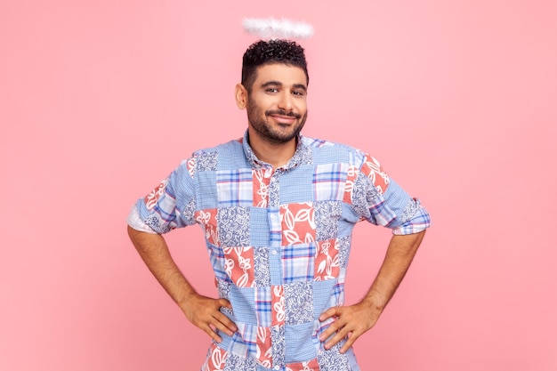 Young adult attractive angelic man with nimb over head wearing blue shirt, standing with hands on hips and looking smiling at camera. Indoor studio shot isolated on pink background.