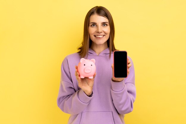 Young adult attractive woman holding piggy bank and smartphone with blank screen for promotion, looking smiling at camera, wearing purple hoodie. Indoor studio shot isolated on yellow background.