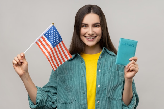 Young adult attractive woman holding united states flag and passport being happy to move abroad to USA wearing casual style jacket Indoor studio shot isolated on gray background