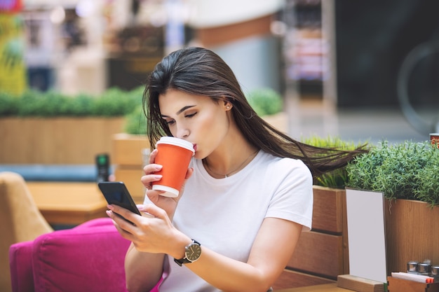 젊은 성인 아름 다운 갈색 머리 소녀 커피와 휴대 전화와 함께 카페에 앉아 행복