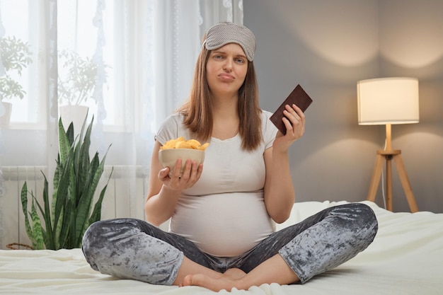 Young adult Caucasian pregnant disappointed woman sitting on bed holding unhealthy junk food snack in hands trying to avoid overeating with chips and chocolate