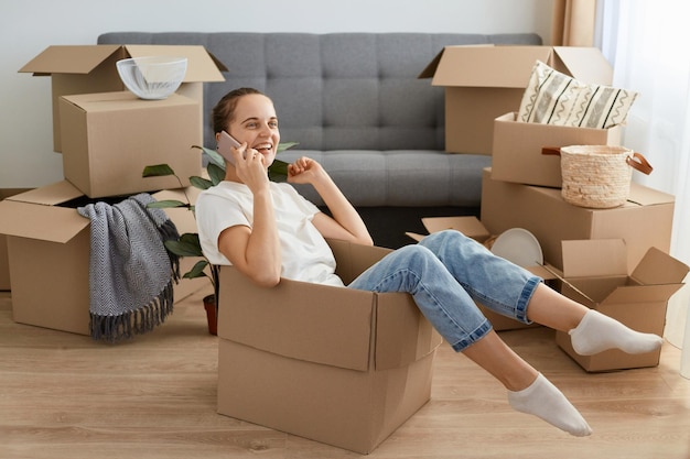 Young adult Caucasian woman wearing white t shirt and jeans sitting in a cardboard box and talking via mobile phone boasting her relocating to a new home with her friend