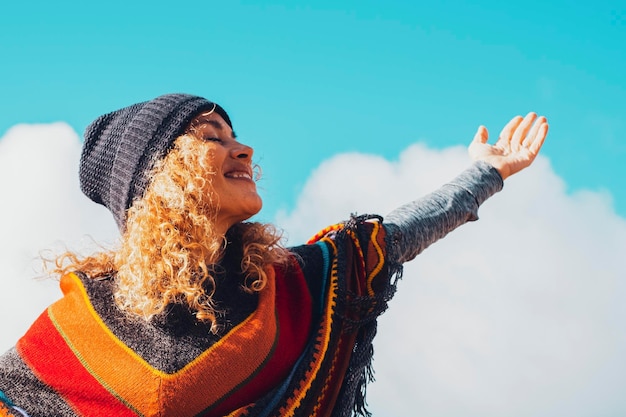 Young adult cheerful lady have fun and enjoy leisure outdoor activity opening arms and smiling against a blue sky background Colorful style poncho and knitted hat for spring winter season
