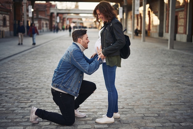 Young adult couple poses happily, pregnant woman. Man looking at the belly