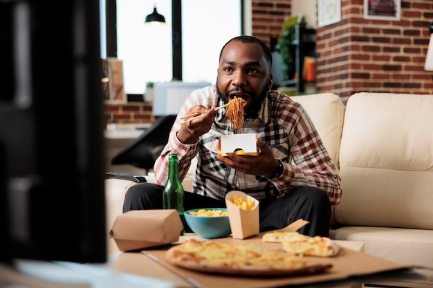 Young adult enjoying noodles package from takeaway delivery, eating food with chopsticks in front of television movie. Watching film on tv channel program, having fun with takeout meal.