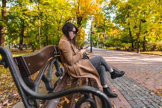 秋の秋のシーズンに都市公園のベンチに座っている若い大人のファッション女性