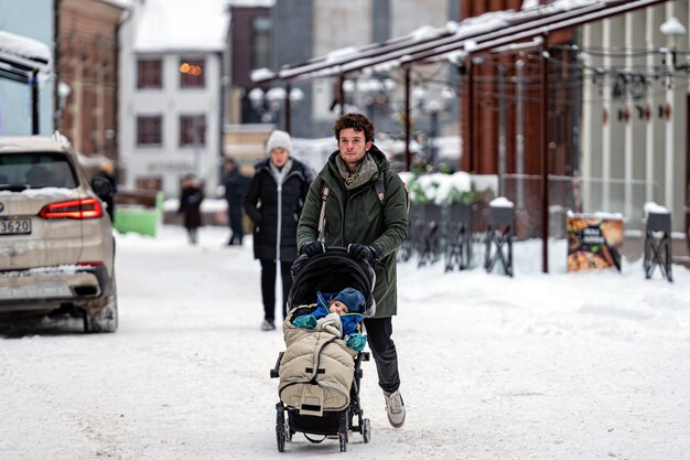 Young adult father pushing baby stroller and walking on snow covered sidewalk at town after blizzard