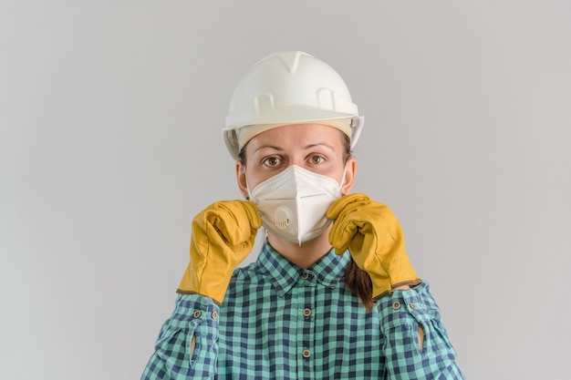 A young adult female construction worker in a protective medical mask stands on a gray background in a white hardhat adjusting a respirator on her face