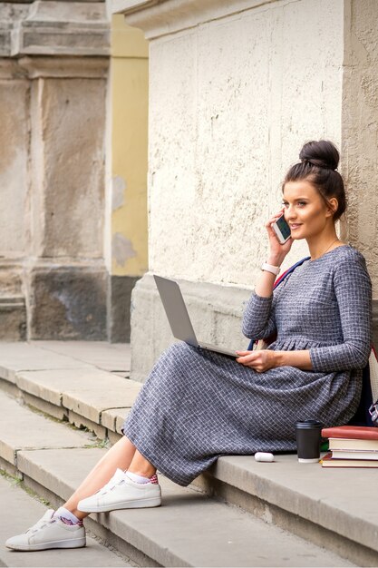 Молодая взрослая студентка разговаривает со смартфоном, сидя на лестнице с ноутбуком возле здания университета