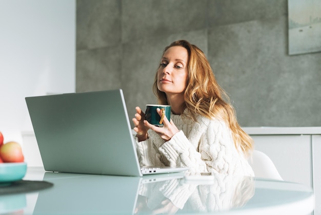 Young adult forty years beautiful blonde woman with long hair working on laptop sitting on the kitchen at home remote work video call freelancer