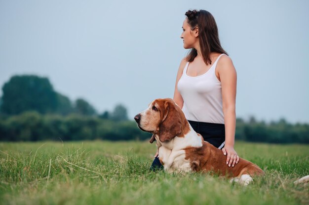 A young adult girl walks with a Basset Hound dog in nature.