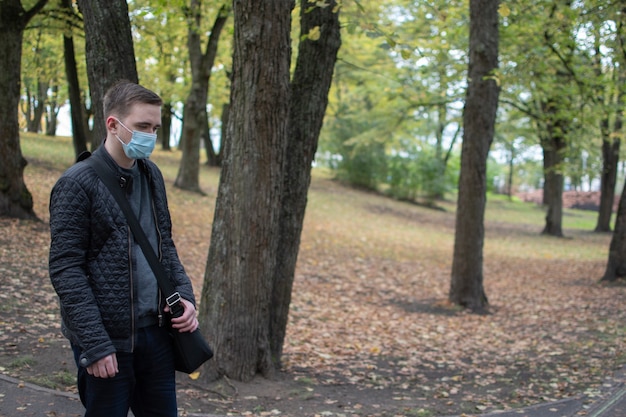 公園のマスクに立っている若い大人の男は、暗い服を着て、緑の公園を思慮深く横に見ています