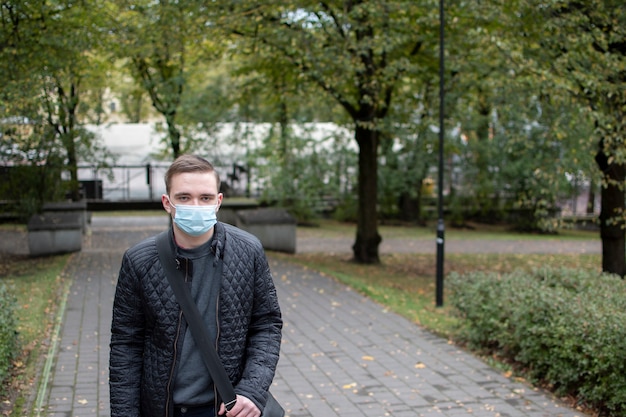 A young adult guy stands in a mask in the park looks to the side thoughtfully, in dark clothes, a green park