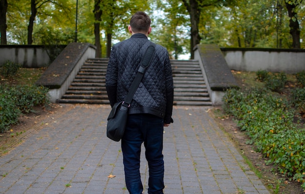A young adult guy stands in the park looking to the side thoughtfully, in dark clothes, green park