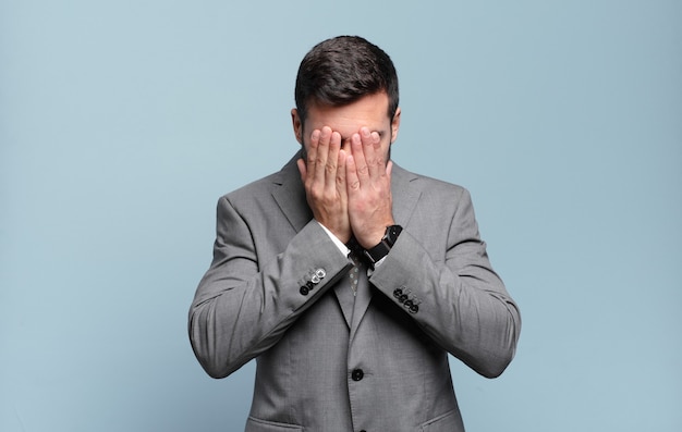 Young adult handsome businessman feeling sad, frustrated, nervous and depressed, covering face with both hands, crying