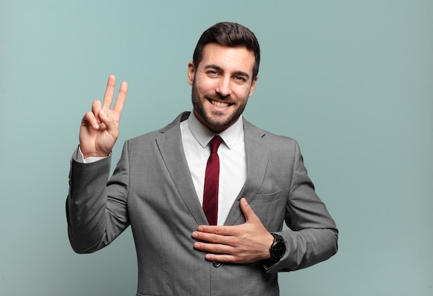 Young adult handsome businessman looking happy, confident and trustworthy, smiling and showing victory sign, with a positive attitude