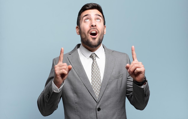 Young adult handsome businessman looking shocked, amazed and open mouthed, pointing upwards with both hands to copy space