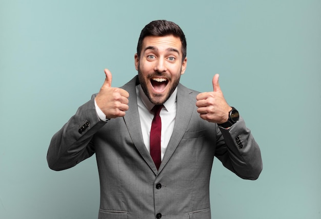 Young adult handsome businessman smiling broadly looking happy, positive, confident and successful, with both thumbs up