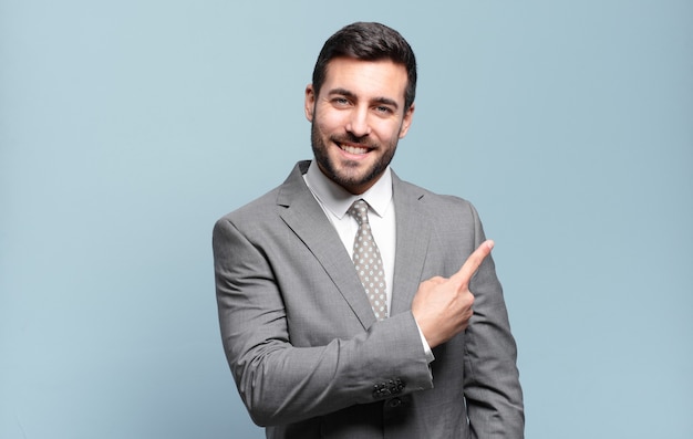 Young adult handsome businessman smiling cheerfully, feeling happy and pointing to the side and upwards, showing object in copy space