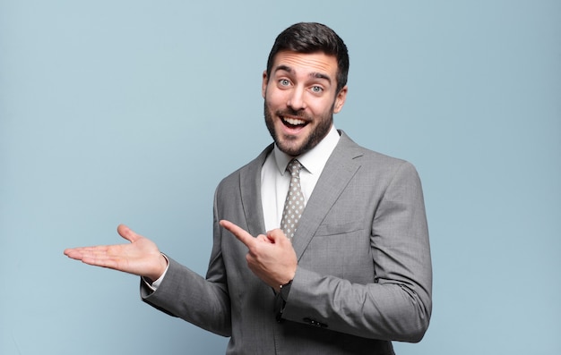 Young adult handsome businessman smiling, feeling happy, carefree and satisfied, pointing to concept or idea on copy space on the side