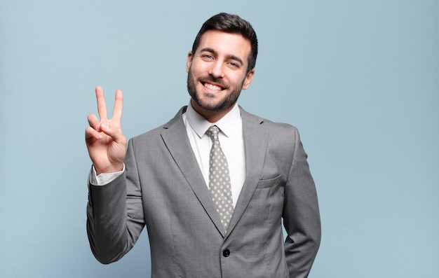 Young adult handsome businessman smiling and looking happy, carefree and positive, gesturing victory or peace with one hand