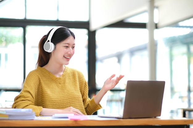 Young adult happy smiling Hispanic Asian student wearing headphones talking on online chat meeting using laptop in university campus or at virtual office College female student learning remotely