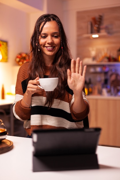 Young adult holding cup of tea while waving on online chatting