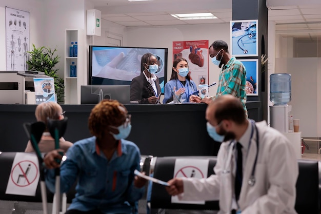 Young adult at hospital reception talking to medical team, discussing about checkup appointment with nurse and receptionist. Male patient in waiting room area during covid 19 pandemic.