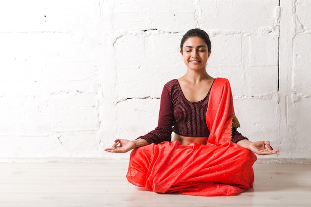 Photo young adult indian woman in sari meditating yoga lotus pose zen like with ok sign mudra gesture at home indoor.