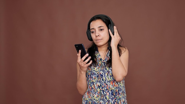 Young adult listening to radio music on wireless headset, playing mp3 song on smartphone app. Listen to melody rhythm and audio sound on headphones, dancing over brown background.