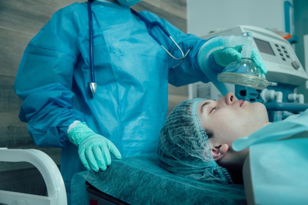 Young adult lying on a medical table in the operating room and a professional doctor holding an anesthetic mask above his face
