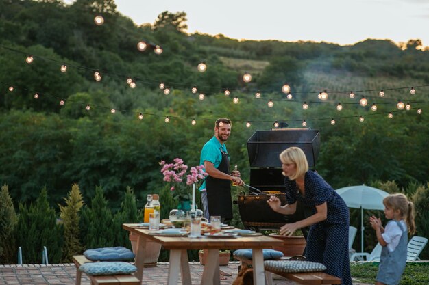 Young adult man barbecuing while his wife and daughter arranging table