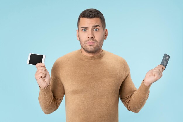 Young adult man holding credit card and smartphone being unsure standing over blue background