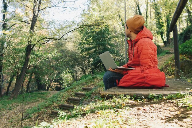 Young adult man in red coat and backpack works with his laptop
in an outdoor forest d