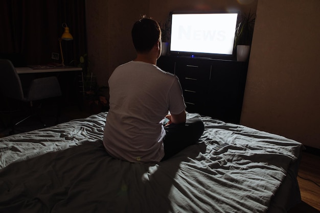 Young adult man sitting on bed looking tv white screen copy\
space night time