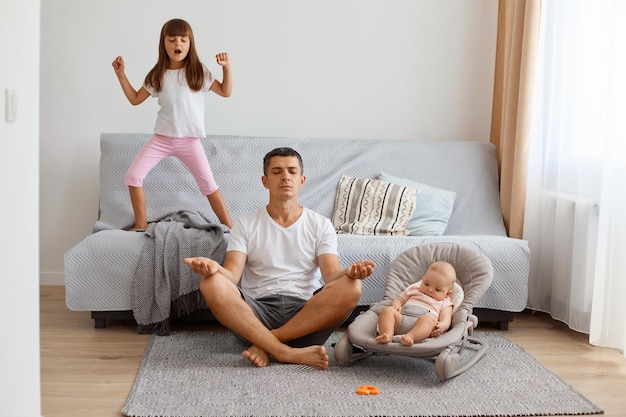 Молодой взрослый человек сидит на полу возле дивана со своим маленьким ребенком в белой футболке и коротких джинсах, пытаясь расслабиться с помощью йоги, в то время как старшая дочь громко поет.