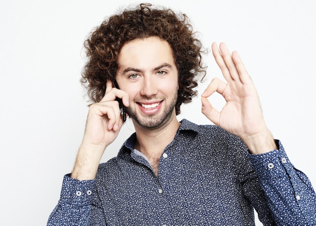 Young adult man talking on the phone standing over white background doing ok sign with fingers