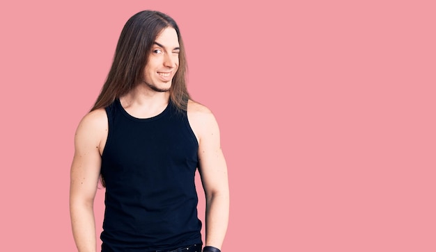 Young adult man with long hair wearing goth style with black clothes winking looking at the camera with sexy expression, cheerful and happy face.
