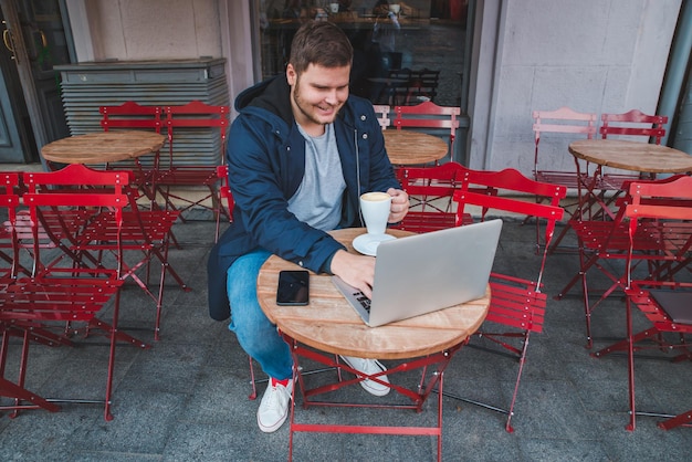 l のカップと屋外カフェでラップトップに取り組んでいる若い成人男性