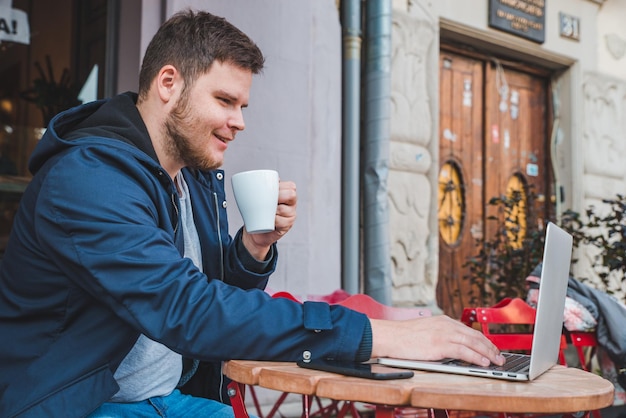 ラテのカップと屋外カフェでラップトップに取り組んでいる若い成人男性