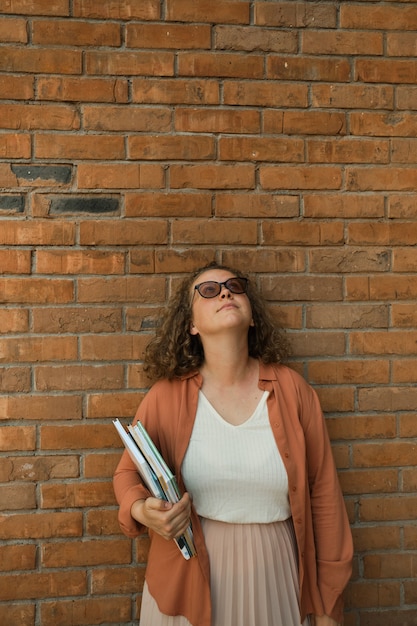 Young adult millennial with curly hair looks to sky Red brick wall Copy space Student holds book