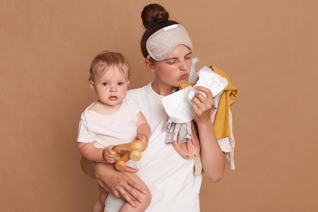 Young adult mother wearing white t shirt and sleeping mask standing isolated over brown background holding baby in hands and posing with lots of kid's clothing smelling diaper bad odor