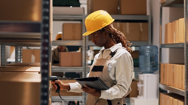 Young adult in overalls checking list of products on tablet, using scanner to count number of merchandise boxes and packages. Female employee working in storage room with goods.