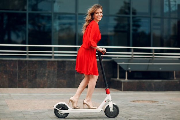 Young adult perfect woman riding electric scooter in red dress at the city