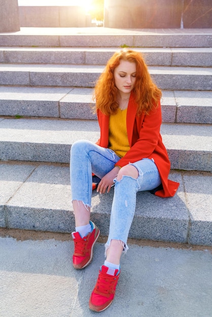 Young adult redhead woman in red coat blue jeans and red sneakers sits