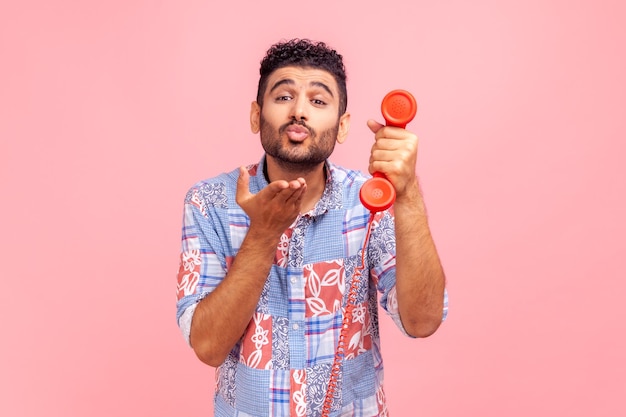 Young adult romantic man call operator wearing blue casual style shirt holding red landline phone and sending air kissing to camera. Indoor studio shot isolated on pink background.