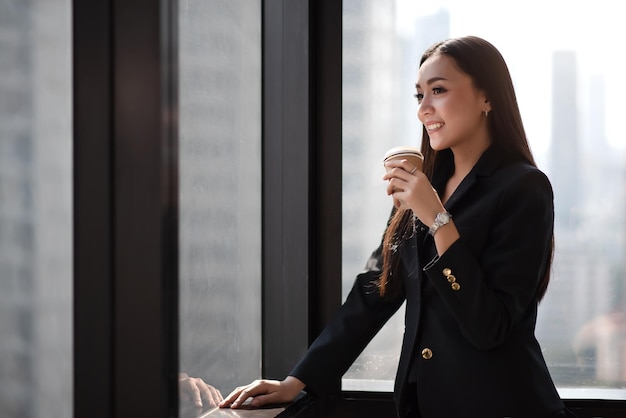 Young adult smart asian business woman in black casual suit holding coffee cup in break time