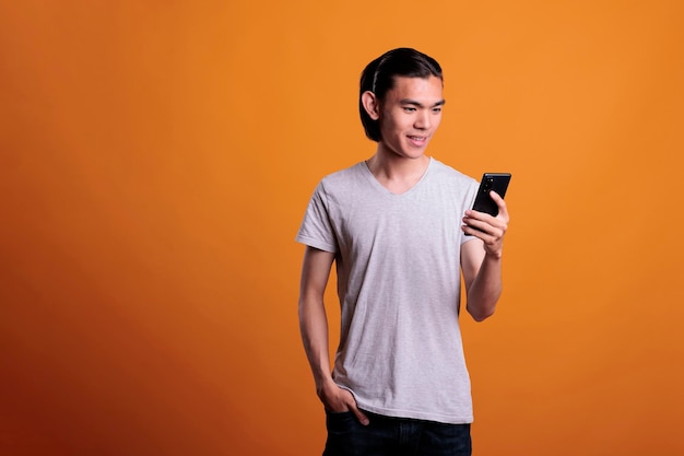 Young adult smiling asian man texting on smartphone, online communication concept. Cheerful teenager standing with hand in pocket, browsing internet on mobile phone, using social media